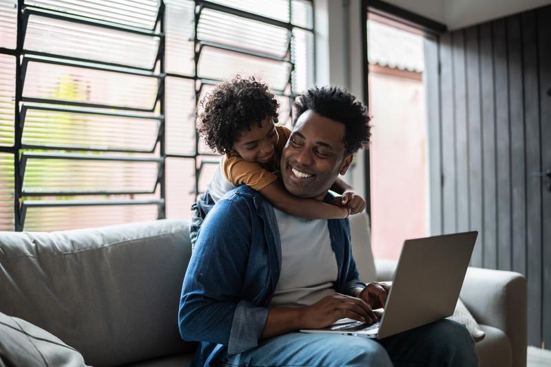 Father sitting on the couch with his son on his back and working on a laptop