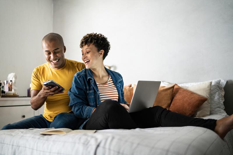 man and woman sitting on bed looking at phone with laptop.