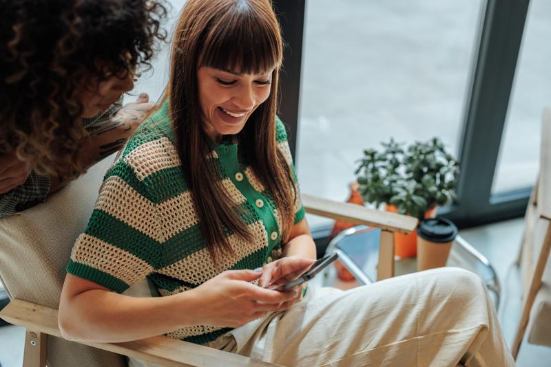 two young women using mobile phone and smiling in a cafe.