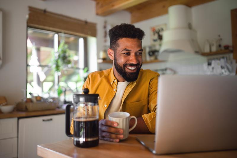 man in kitchen with coffee cup in hand looking at laptop screen.