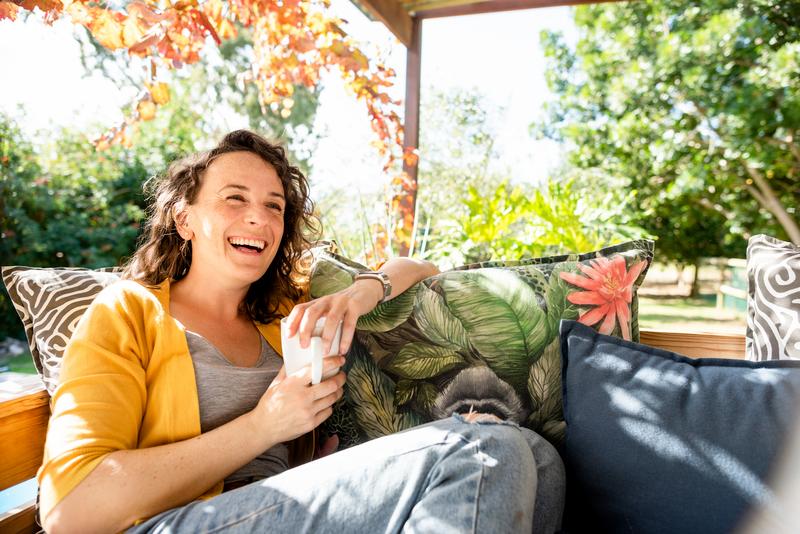 young woman laughing while relaxing outside on a sofa on her patio and drinking a cup of coffee.