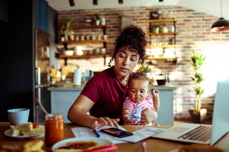 Image: woman sitting at table holding infant and using calculator .