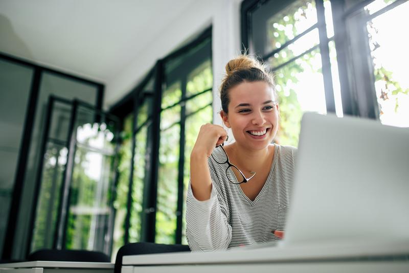 Image: woman holding eyeglasses smiling working on laptop.