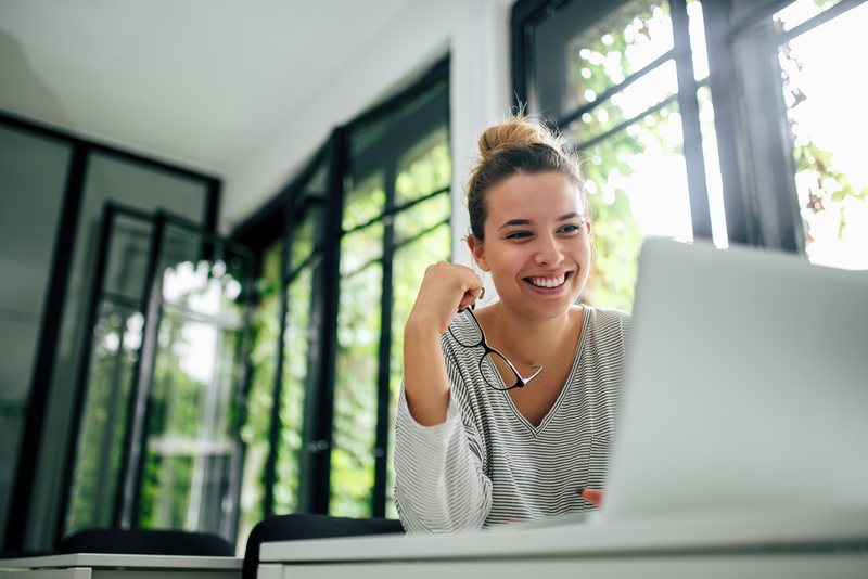 Woman holding eyeglasses smiling working on laptop
