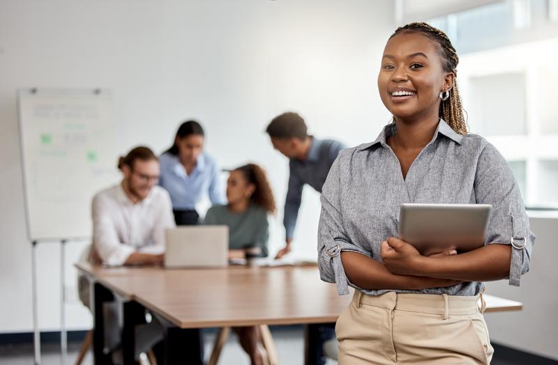 Image: woman with tablet in business setting with colleagues in background.
